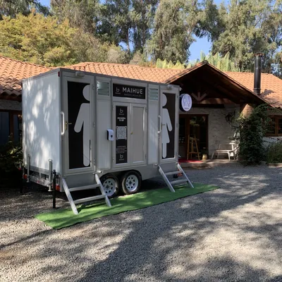 Tráiler de baños móviles Maihue instalado sobre césped frente a una casa de campo con techo de tejas