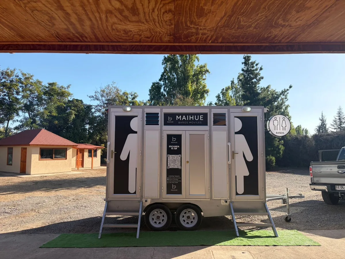 Tráiler de baños móviles premium Maihue instalado bajo una pérgola de madera en una parcela, sobre una alfombra de pasto y rodeado de pinos