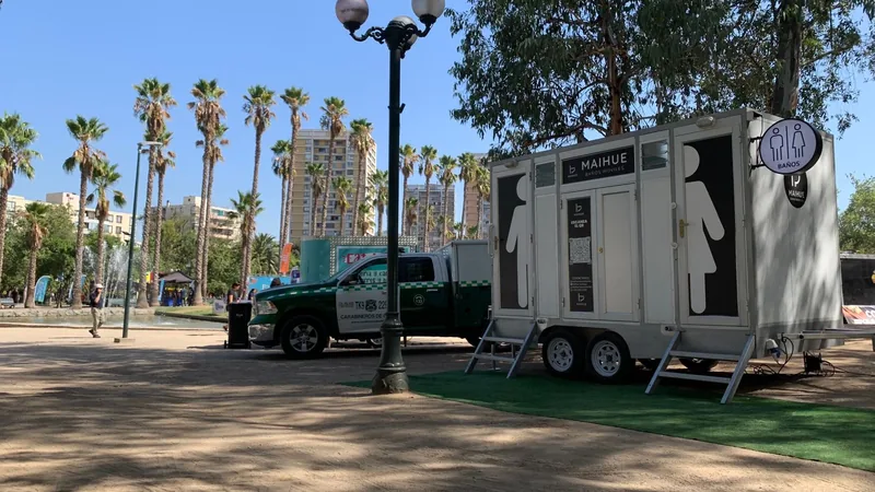 Tráiler de baños móviles premium Maihue instalado sobre césped en una plaza de Santiago con palmeras, junto a una camioneta municipal